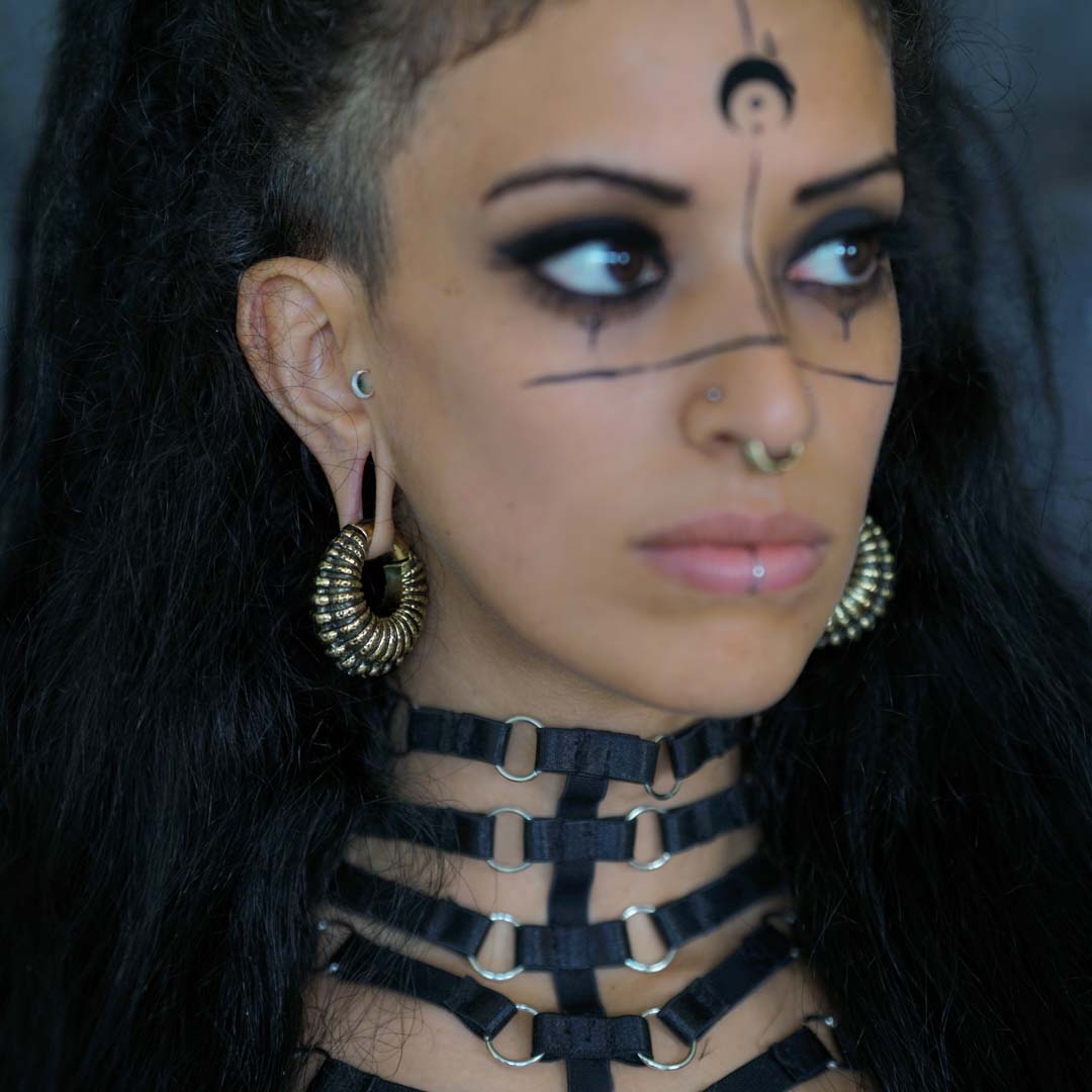 A person with long dark hair and a side undercut is shown. They are wearing Kaya Movement's TRILOBITE Large Biomechanical Clicker Ear Weights in Gold Brass, sized at 0 gauge, tribal gauge earrings, a septum piercing, lip piercing, and dark makeup with black lines and symbols on their face. They are also wearing a black harness necklace with metal rings.